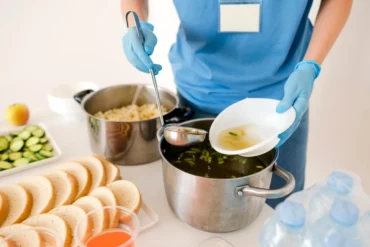 A food service worker in blue gloves ladles hot vegetable soup into a disposable bowl, surrounded by neatly arranged bread, cucumbers, and water bottles, emphasizing hygiene and food safety in a shared kitchen setting.