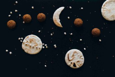 Creative food arrangement on a dark background featuring pita bread, falafel balls, and scattered chickpeas, artistically resembling a celestial scene, representing Arabic cuisine and its global influence, including its arrival in Nepal.